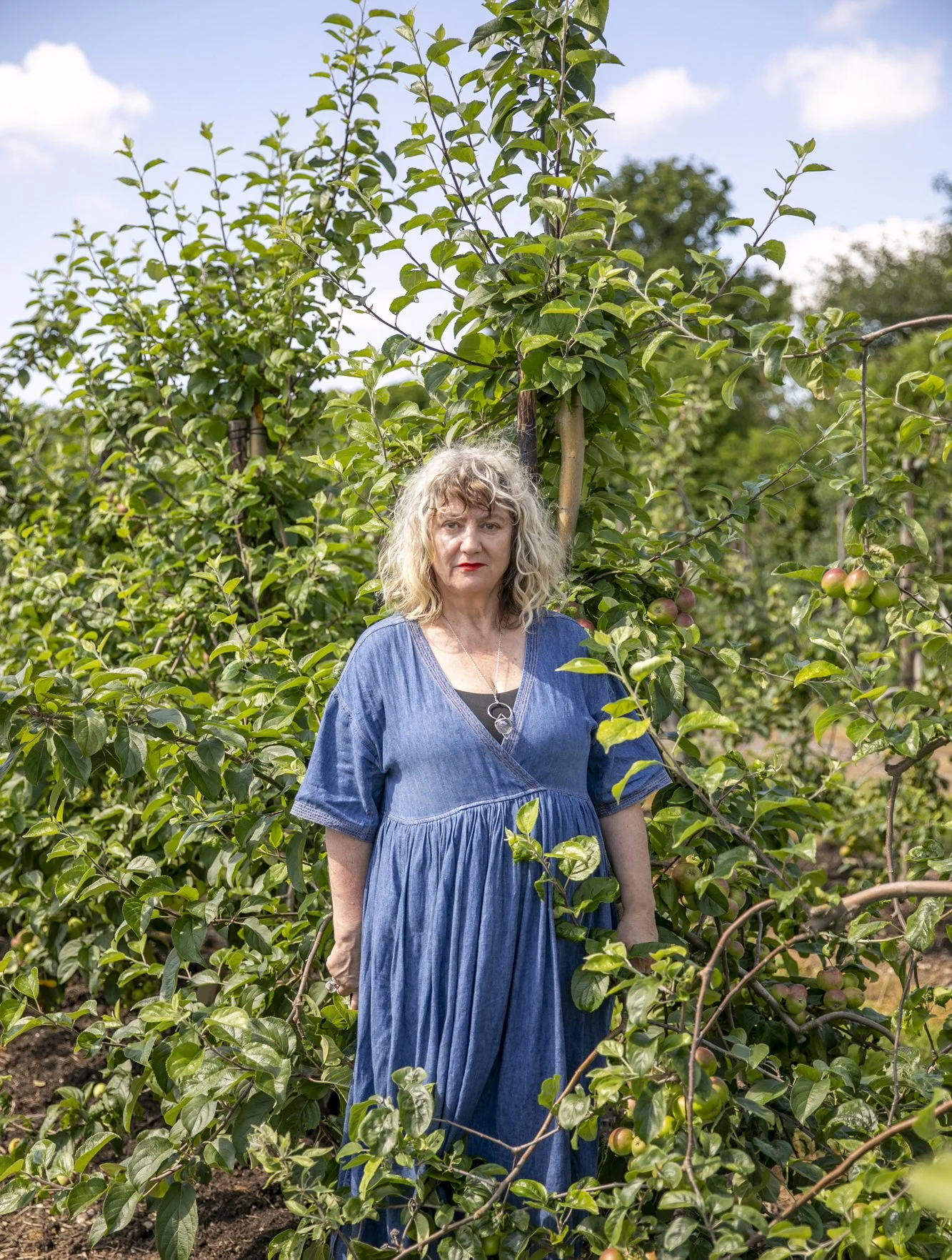 A woman with curly blonde hair wearing a blue dress standing among green apple trees in a garden.