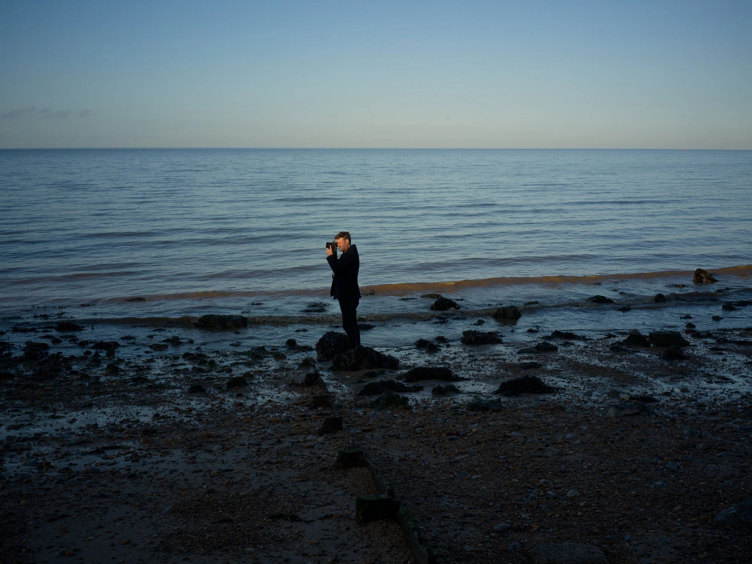 A man in a black coat stands on a rocky beach, taking a photo with his camera of the calm ocean and sky at dusk or dawn.