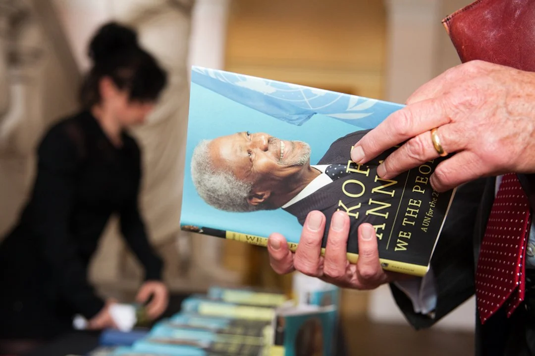 Close-up of a person holding a book titled "We the People" with a photograph of an older man with gray hair and a suit on the cover. A woman is in the background at a table with more books, out of focus.