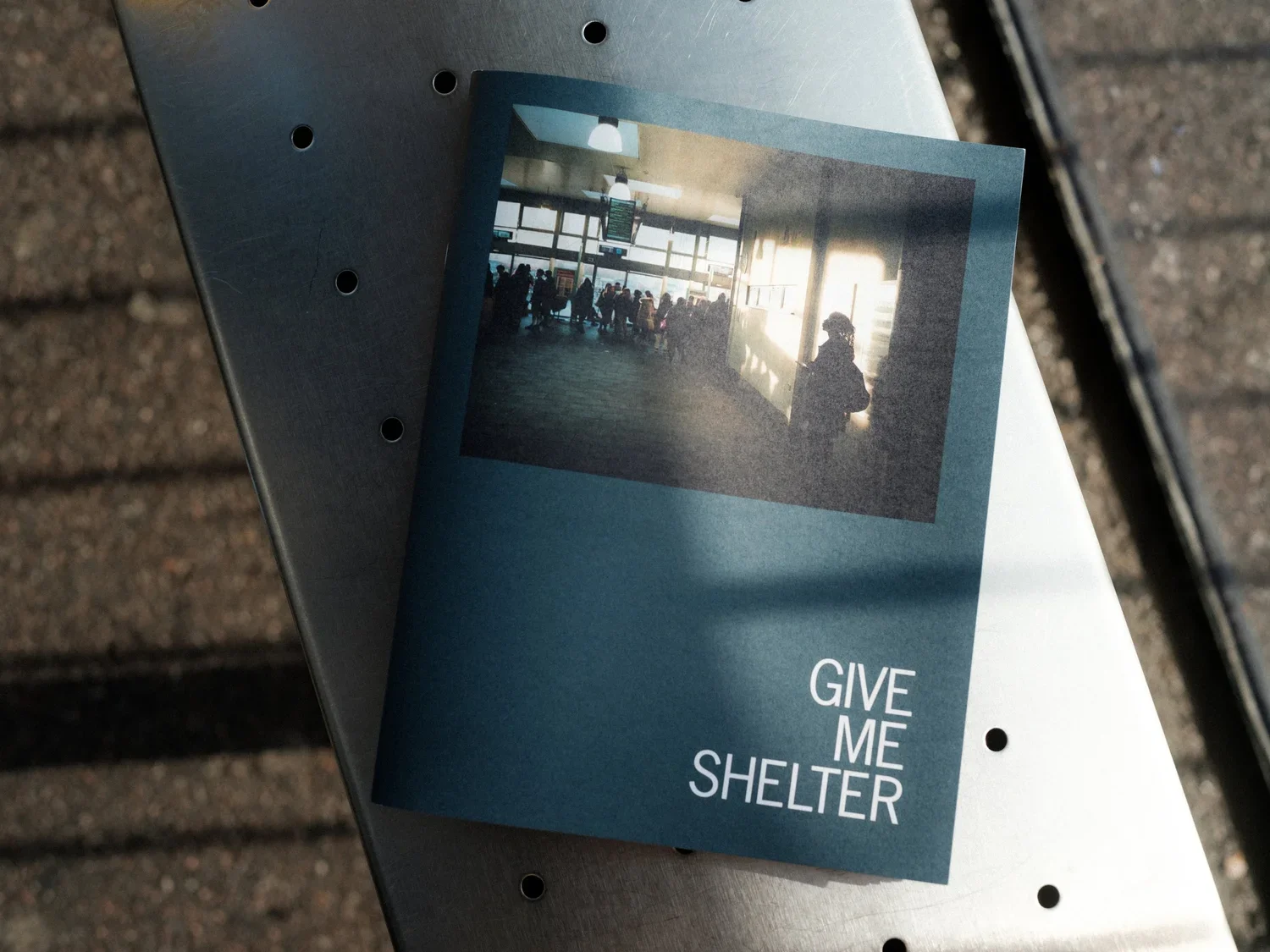 A pamphlet with the words 'Give Me Shelter' and a photo of an airport terminal with people waiting and sunlight streaming through windows, placed on a gray metal surface outside.