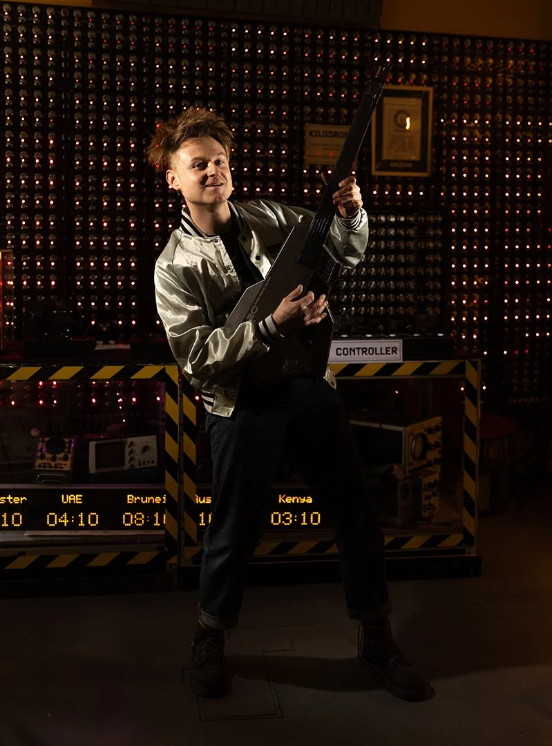 Look Mum No Computer (aka Sam Battle) with spiked hair playing a black guitar in a dimly lit room with a wall of small red lights and digital screens displaying flight information.