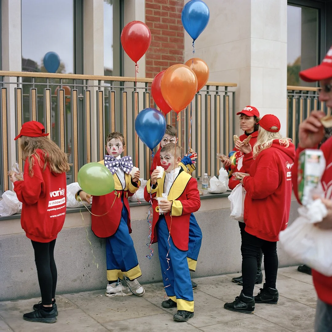 Children and adults dressed as clowns and in red jackets, some holding colorful balloons, standing outside near a railing and a building, eating snacks.