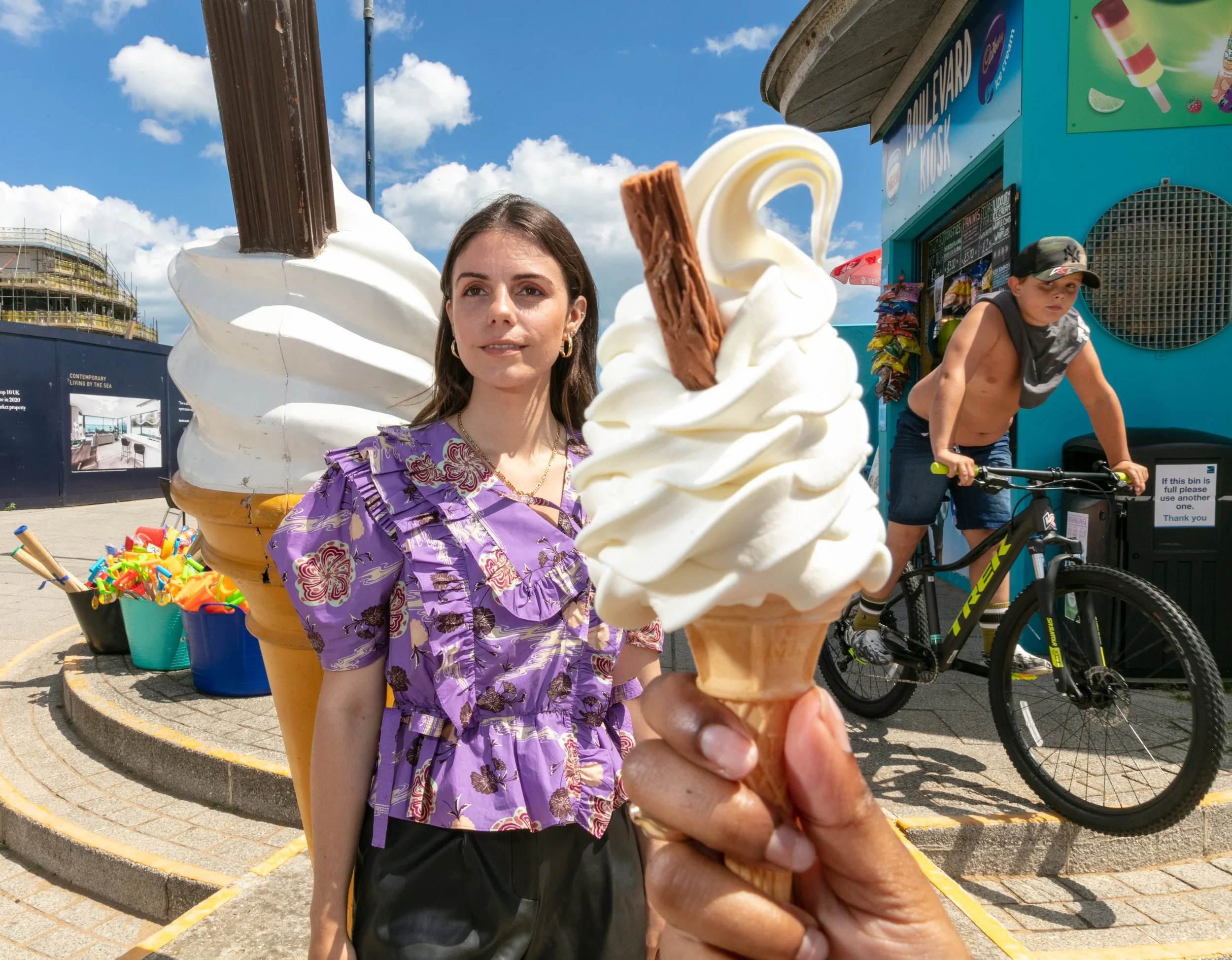 A person holding an ice cream cone topped with vanilla ice cream, a chocolate, and a wafer. A woman in a purple floral dress stands behind, near a large ice cream sculpture and a blue popcorn vending machine outdoors against a bright blue sky with wh
