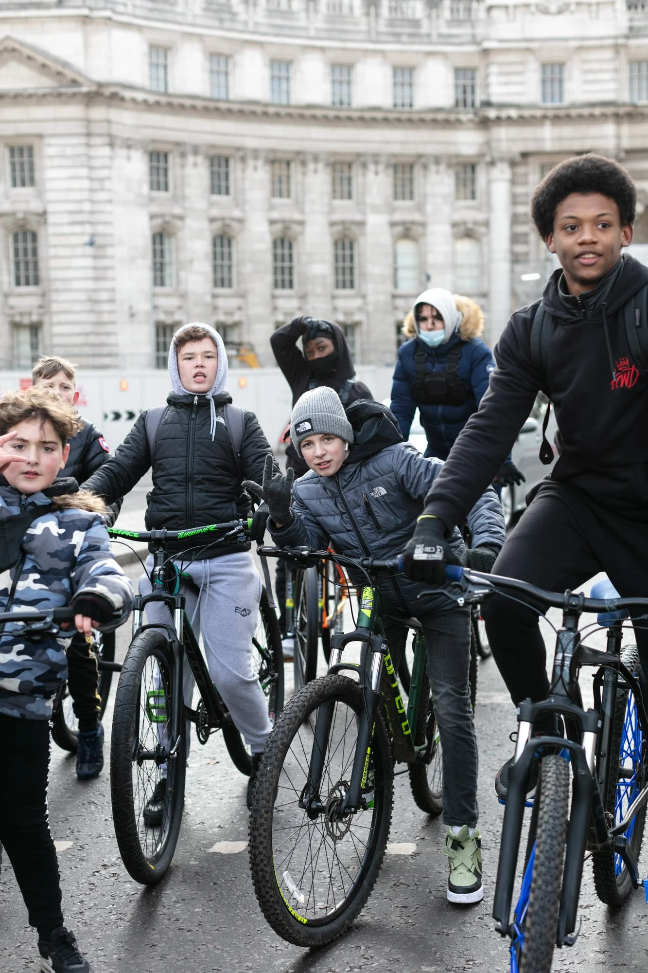 A group of children and teenagers with bicycles gathering outdoors in front of a large historic building. They are wearing warm clothing, and some are making peace signs or greeting gestures.