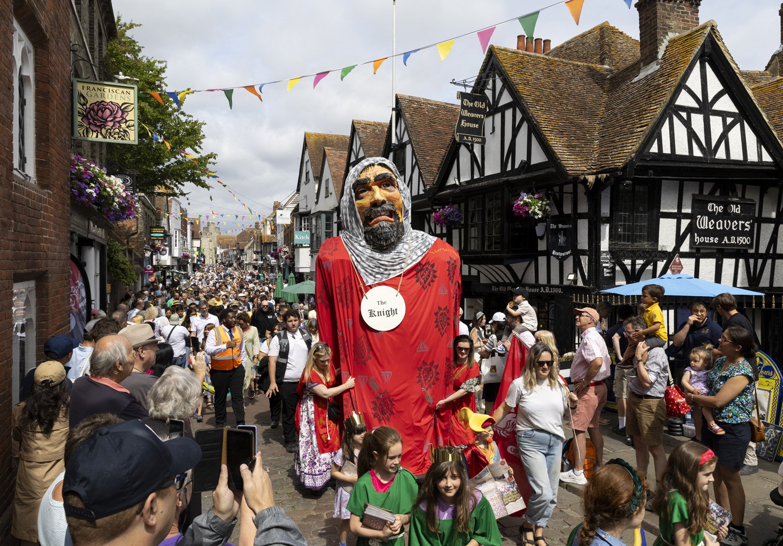 Crowd of people gathered in a street during a festival with a large puppet of a man labeled 'The Knight' in the center. The street is decorated with colorful flags and traditional-style black-and-white buildings.
