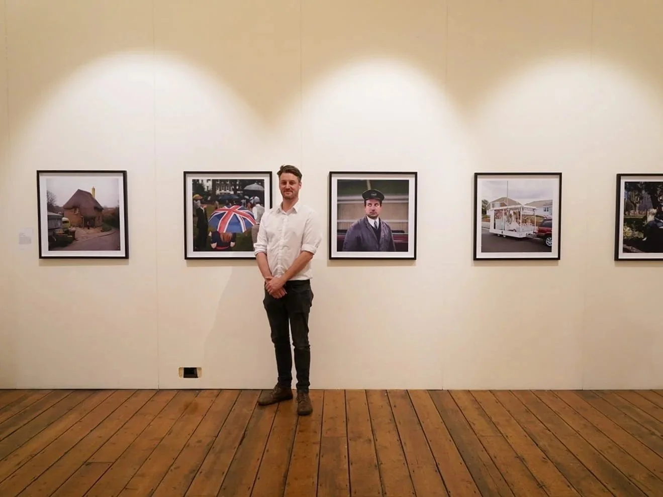 A man standing in an art gallery in front of framed photographs on a beige wall, with wooden flooring and ceiling lights.