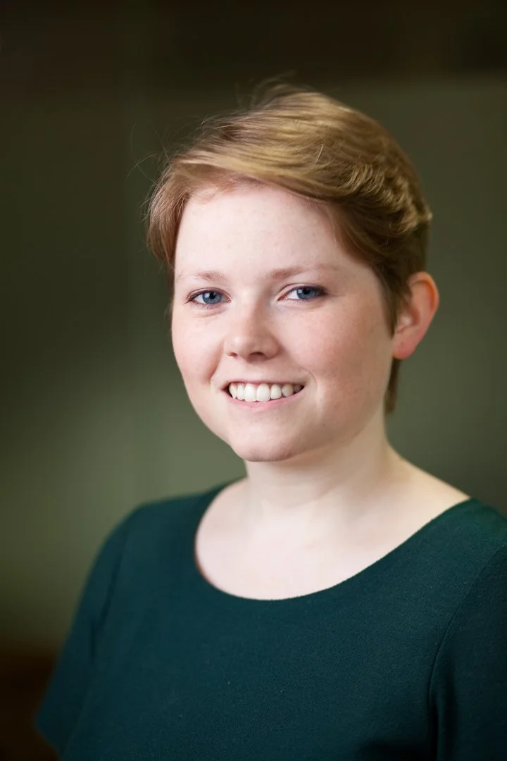 A young woman with short light brown hair and blue eyes smiling, wearing a dark green top, against a blurred green background.