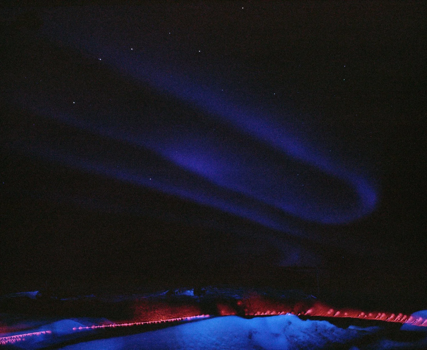 Night sky with visible stars and glowing aurora borealis over a snow-covered landscape, with faint red lights on the ground.