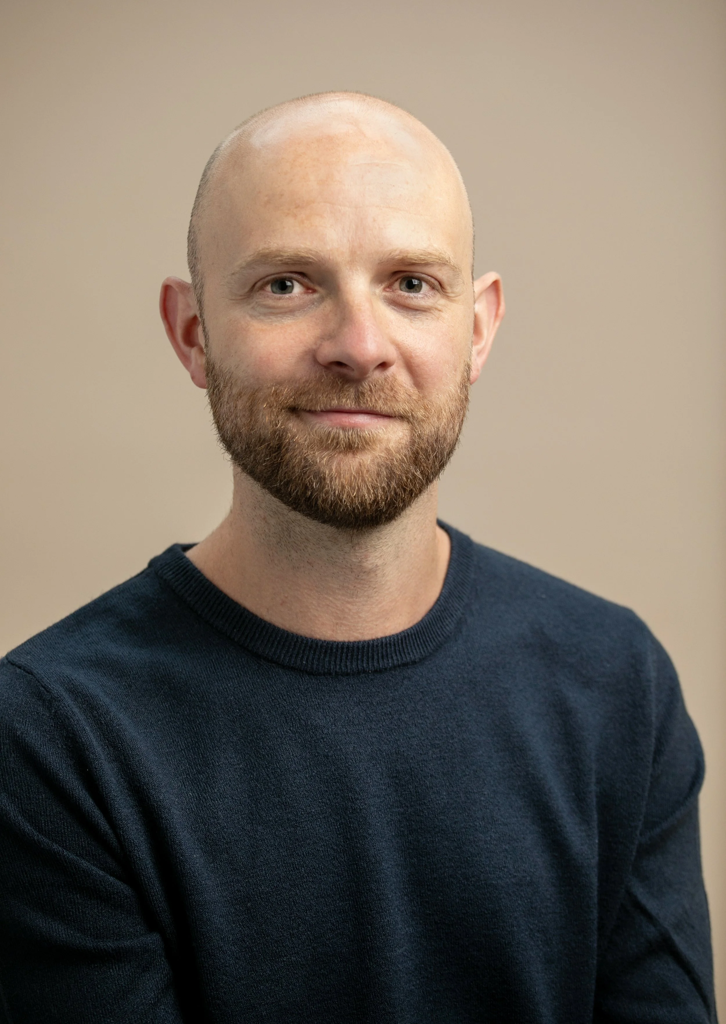 A close-up of a man with a beard and shaved head, wearing a dark navy sweater, standing against a beige background.