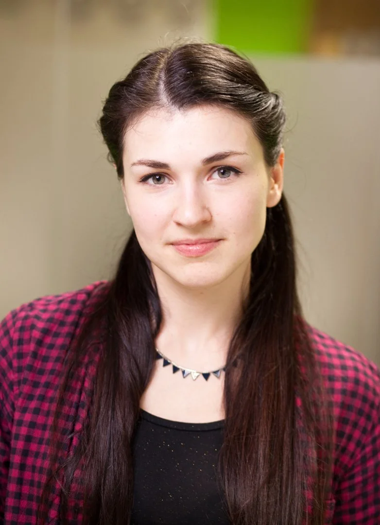 A young woman with long dark hair, wearing a black top and a red plaid blazer, looking at the camera with a slight smile, standing indoors with a blurred neutral background.