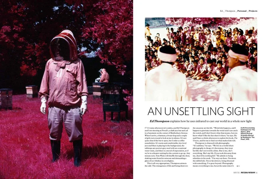 Person wearing a beekeeping suit in a pink surreal landscape with pink trees and grass, with a person sitting on a park bench in the background.