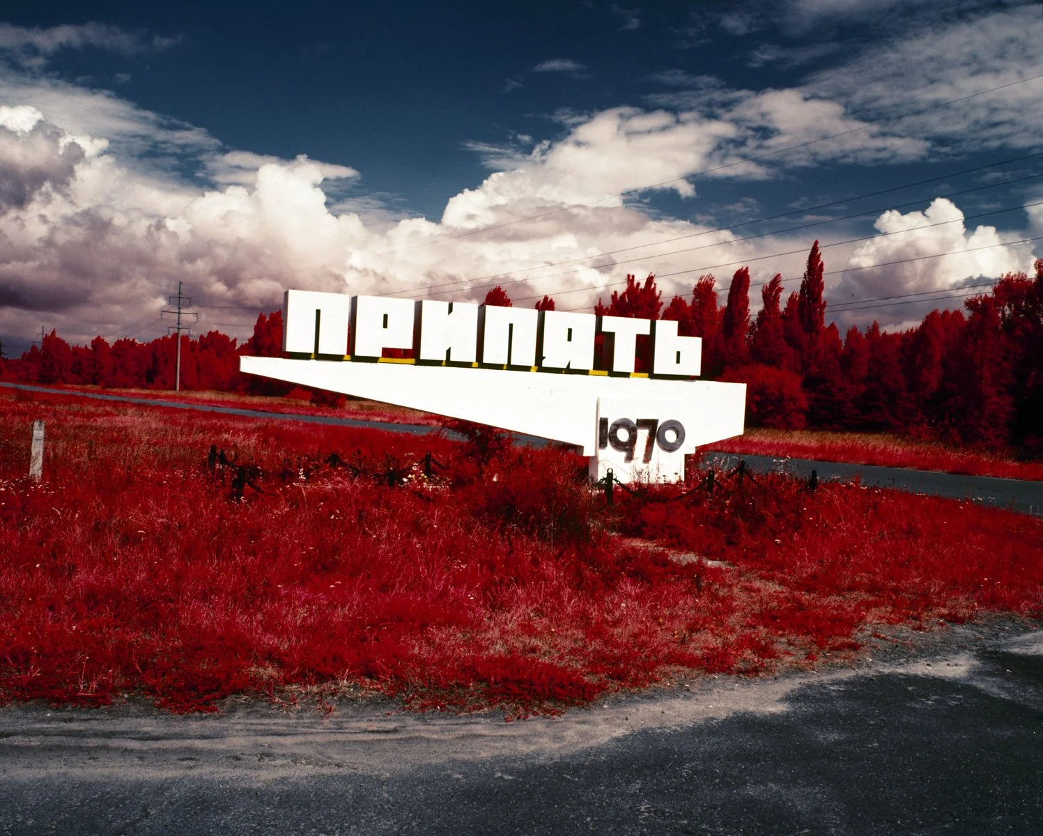 A white roadside sign with the word 'ИРСОНЬ' and the year 1970 written on it, located on a grassy roadside with trees and a cloudy sky in the background.