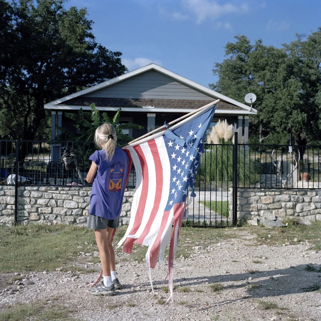 A young girl with blonde hair in a ponytail, wearing a blue T-shirt, gray shorts, and sneakers, stands outdoors near a black metal fence, holding a torn American flag. Behind her, there is a house with gray siding, a satellite dish, trees, and bushes