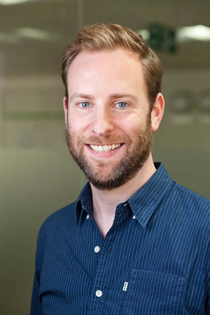 Portrait of a man with light brown hair, blue eyes, and a beard, smiling, wearing a dark blue, pinstriped button-up shirt, indoors with a blurred background.
