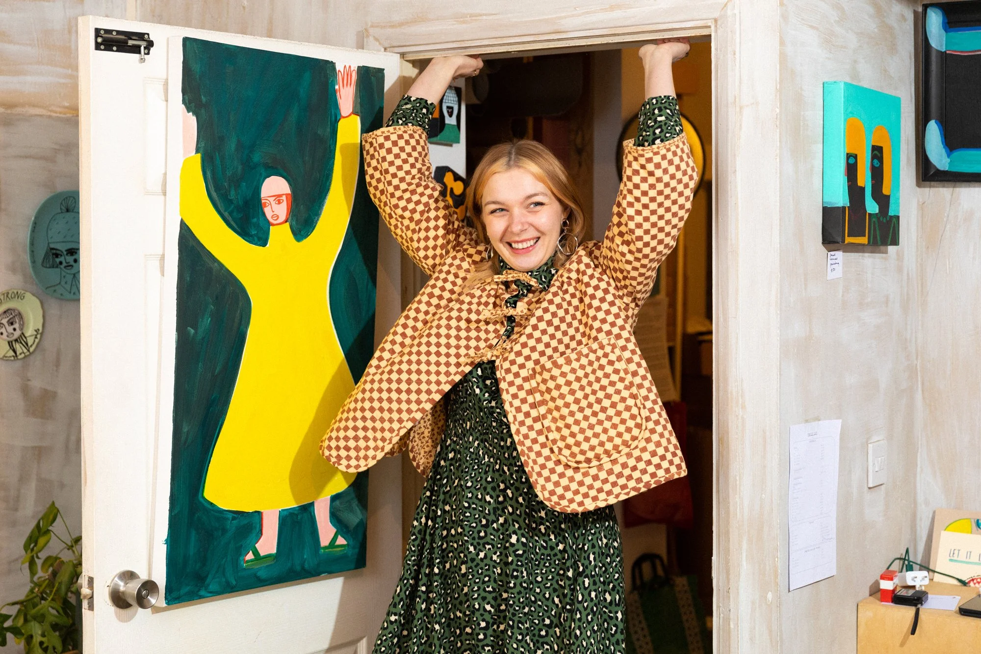 A smiling young woman with blonde hair stands in an art gallery doorway with her arms raised above her head. She wears a patterned brown and black checkered jacket over a black dress with green and white abstract patterns. Behind her, colorful abstra