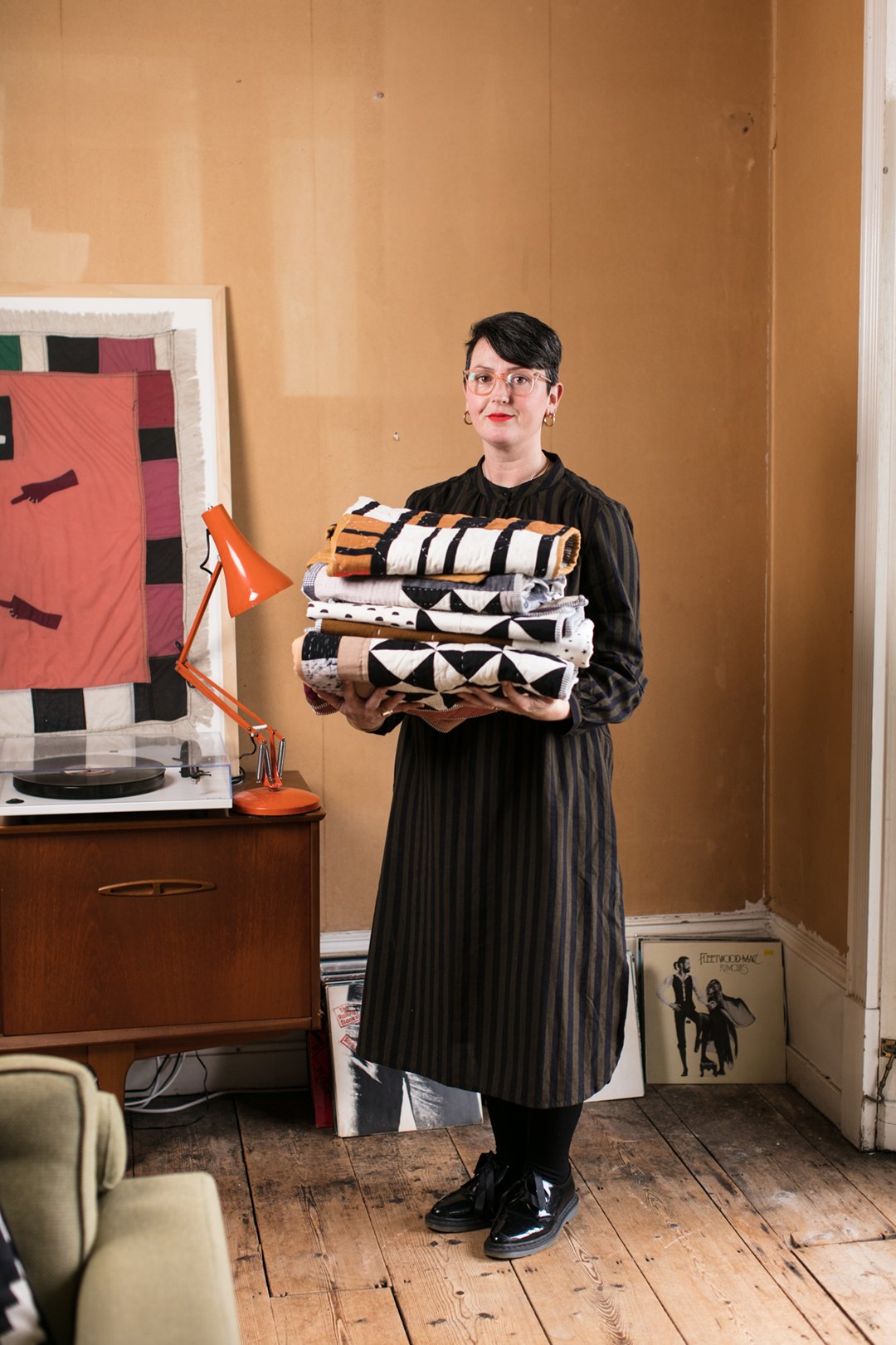 A woman with short dark hair and glasses holding a stack of folded blankets or textiles, standing in a room with wooden floors and a beige wall, next to a wooden cabinet with a vintage orange desk lamp and a record player.
