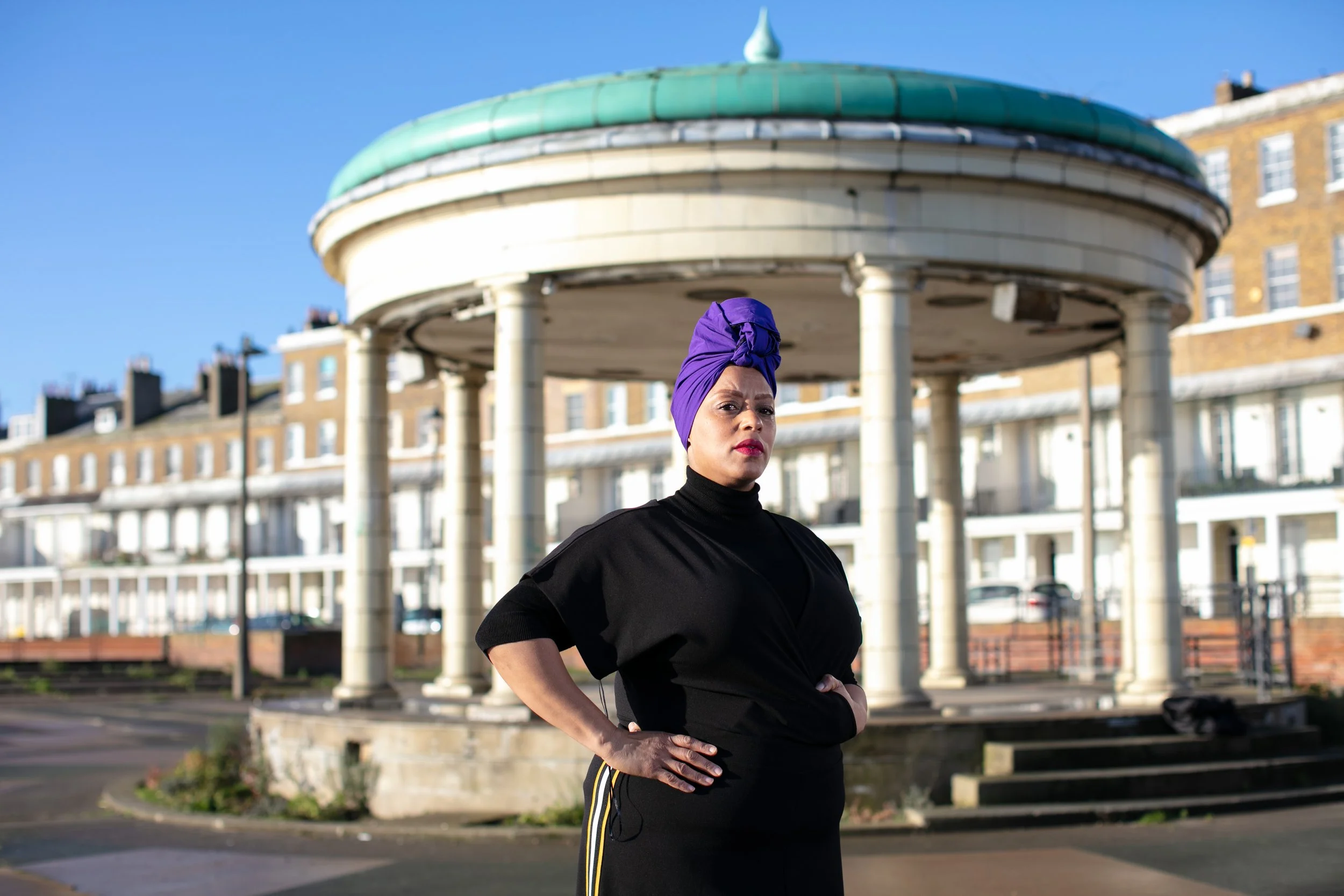 A woman wearing a purple headwrap and black top standing outdoors in front of an ornate gazebo with a green copper roof and brick buildings in the background.