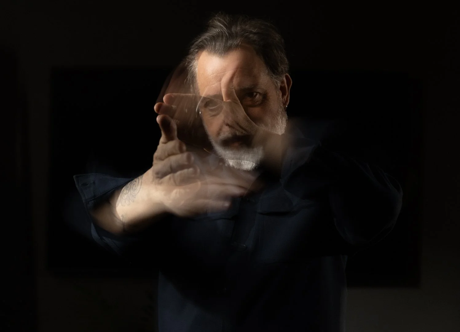 A man with gray hair and a beard, wearing a dark jacket, is holding a glass of drink to his lips, with a mirror image reflecting his face and hand in the dark background.