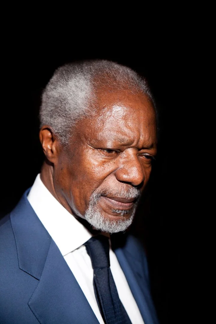 An elderly man with gray hair and a beard wearing a dark blue suit, white shirt, and dark tie, looking down with a serious expression against a black background.