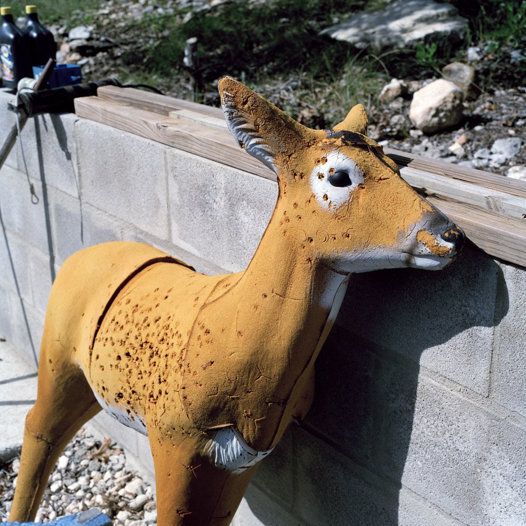 A weathered, vintage deer statue with a yellowish-orange body and white accents, leaning against a concrete block wall outdoors.