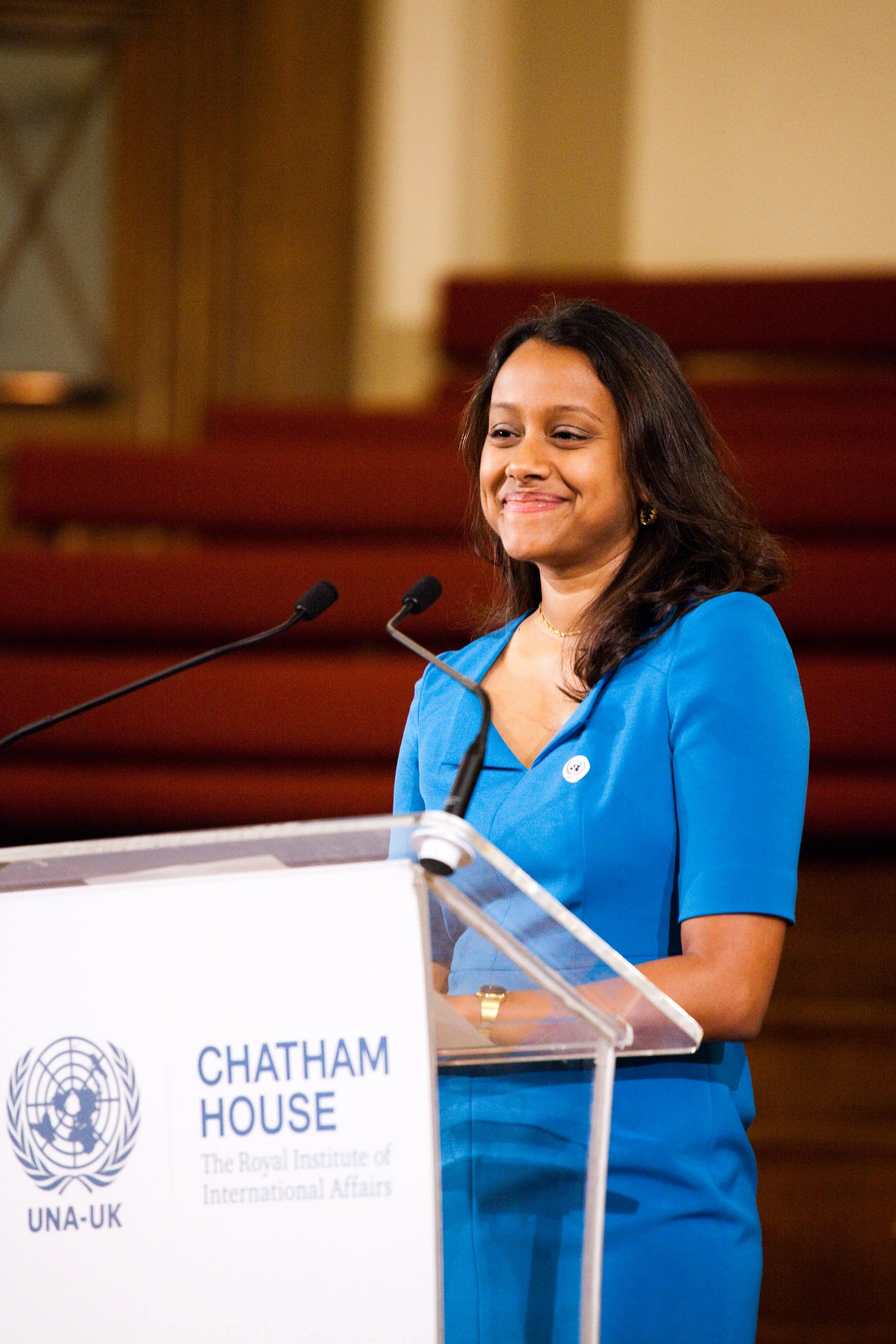A woman in a blue dress smiling at a podium with the United Nations logo, speaking at Chatham House.