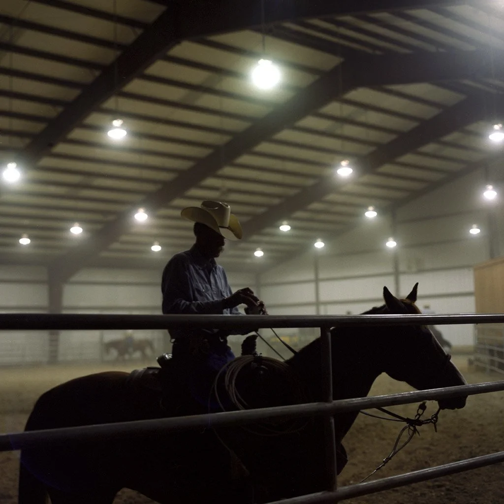 A person wearing a cowboy hat sitting on a horse inside an indoor riding arena, with bright overhead lights and a metal railing.