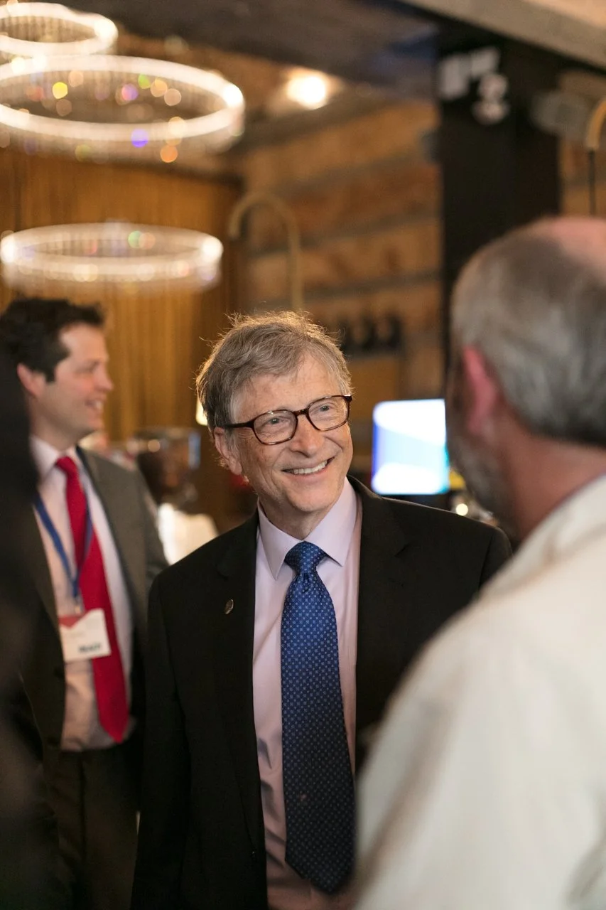 A man wearing glasses and a suit with a blue tie smiling and talking to another person inside a warmly lit room with wooden walls and chandeliers hanging overhead.