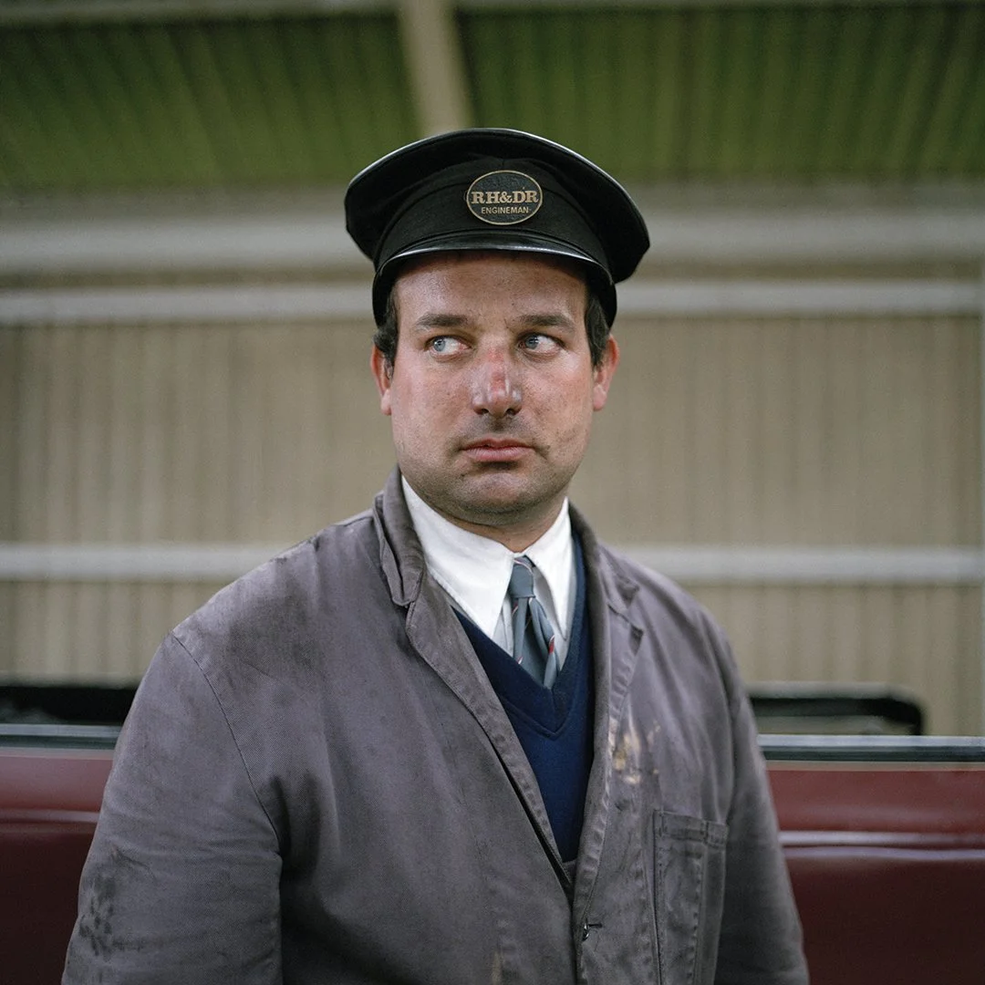 A man wearing a black train conductor hat with a badge that says RH & DR ENGINEMAN, a brown jacket, white shirt, and tie, standing outside near a train.