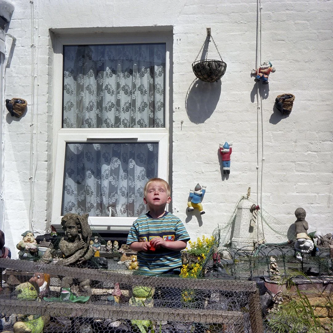 A young boy wearing a striped shirt stands in a garden filled with various garden ornaments and figurines. Behind him is a white brick wall with a window and several decorative items, including hanging planters, small sculptures, and gnomes.