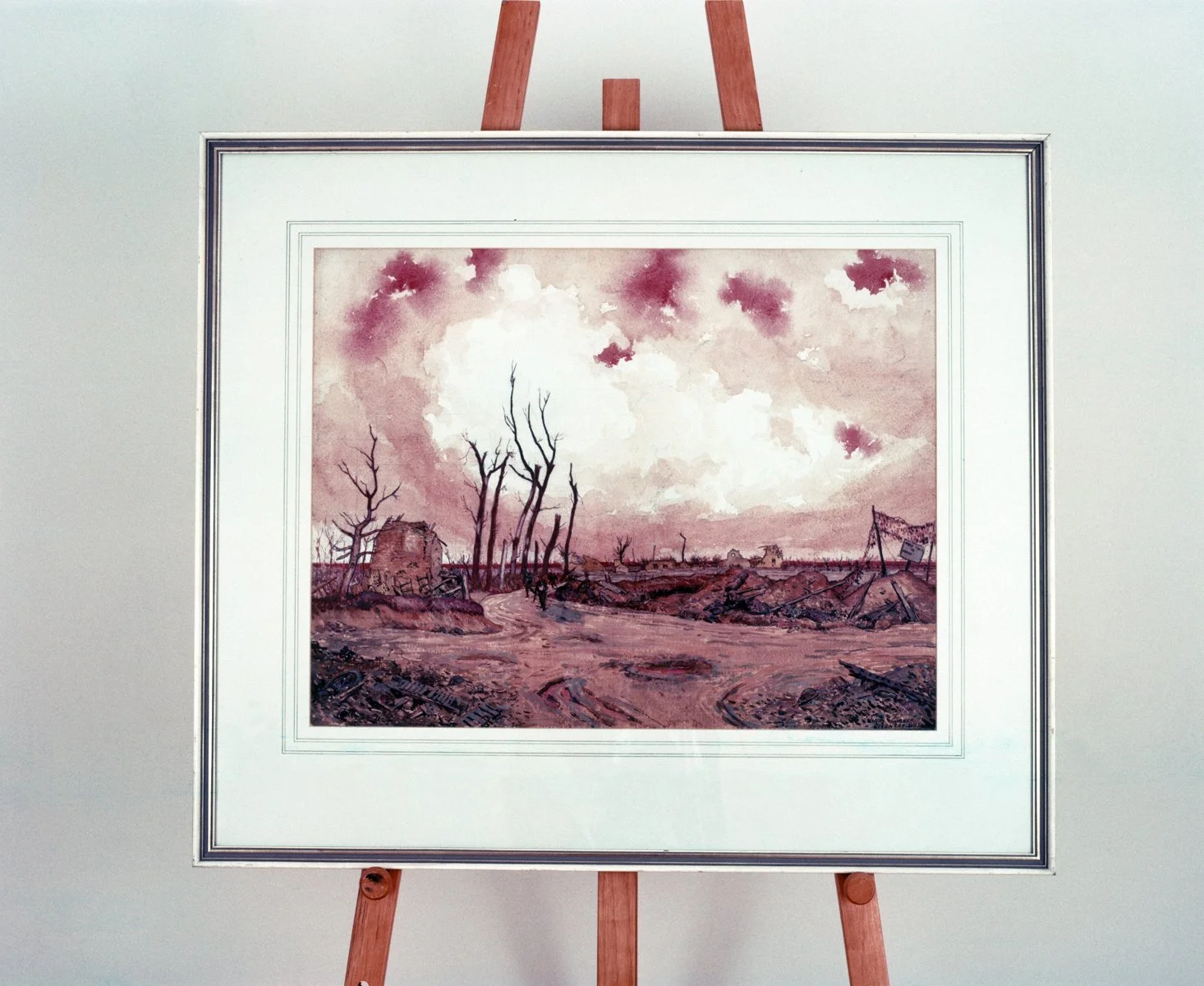 A framed watercolor painting of a desolate rural landscape with dead trees, a dirt road, and a cloudy sky, displayed on an easel.