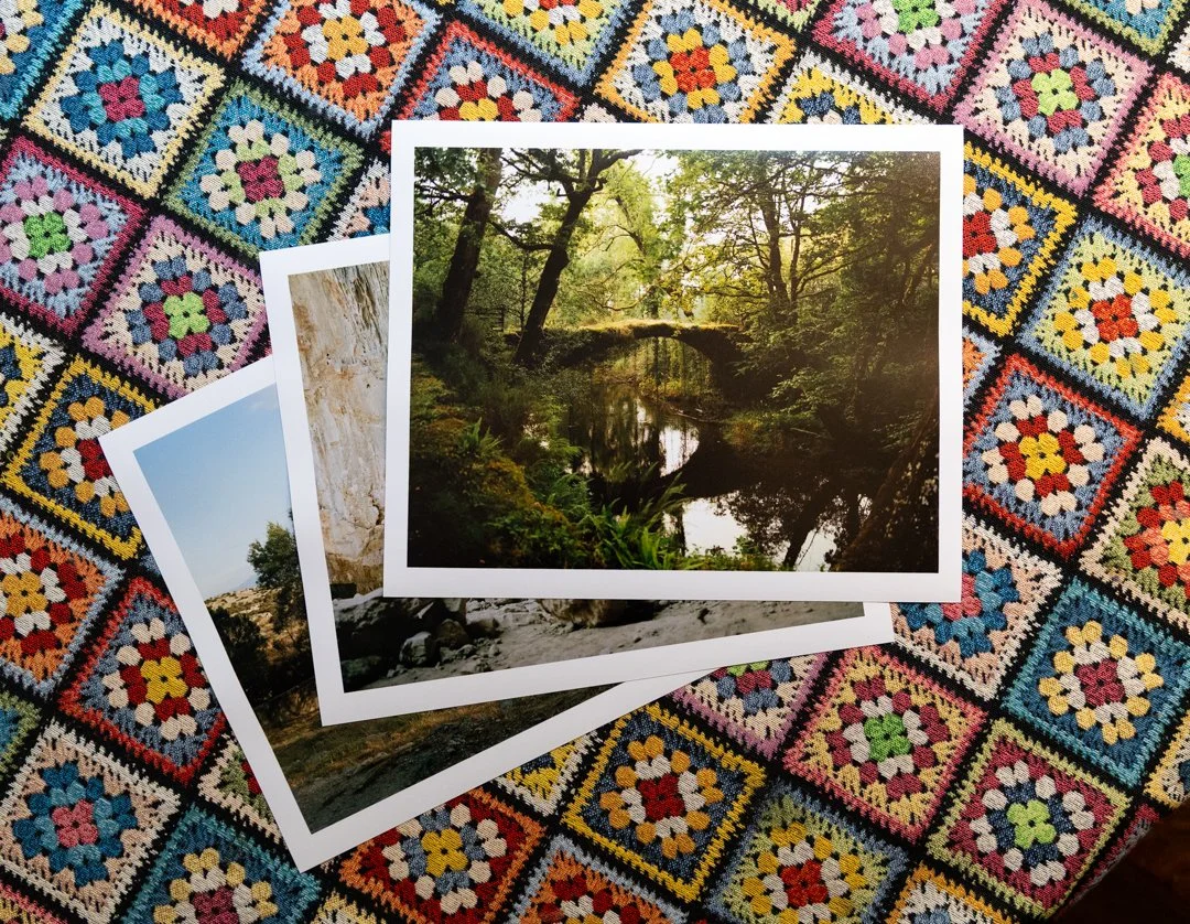 Three printed photographs of nature scenes, including a forest with a river and a stone bridge, and a rocky landscape, lay on a colorful crocheted blanket with floral patterns.