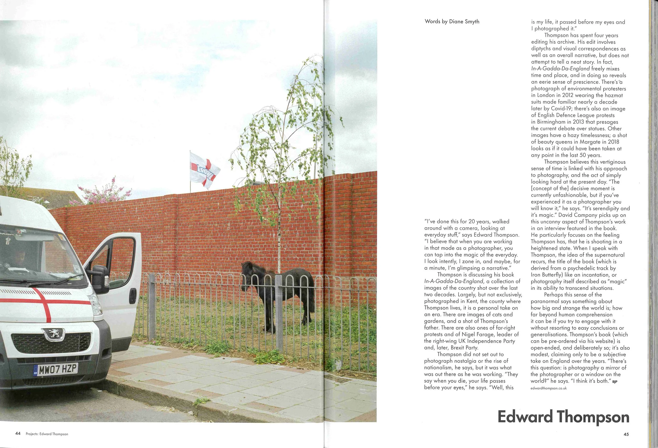 Photograph of a suburban street with white vans parked along the sidewalk, a tall brick wall, a tree with green leaves, pink blossoms, a flag in the background, and a black dog sitting near the fence.