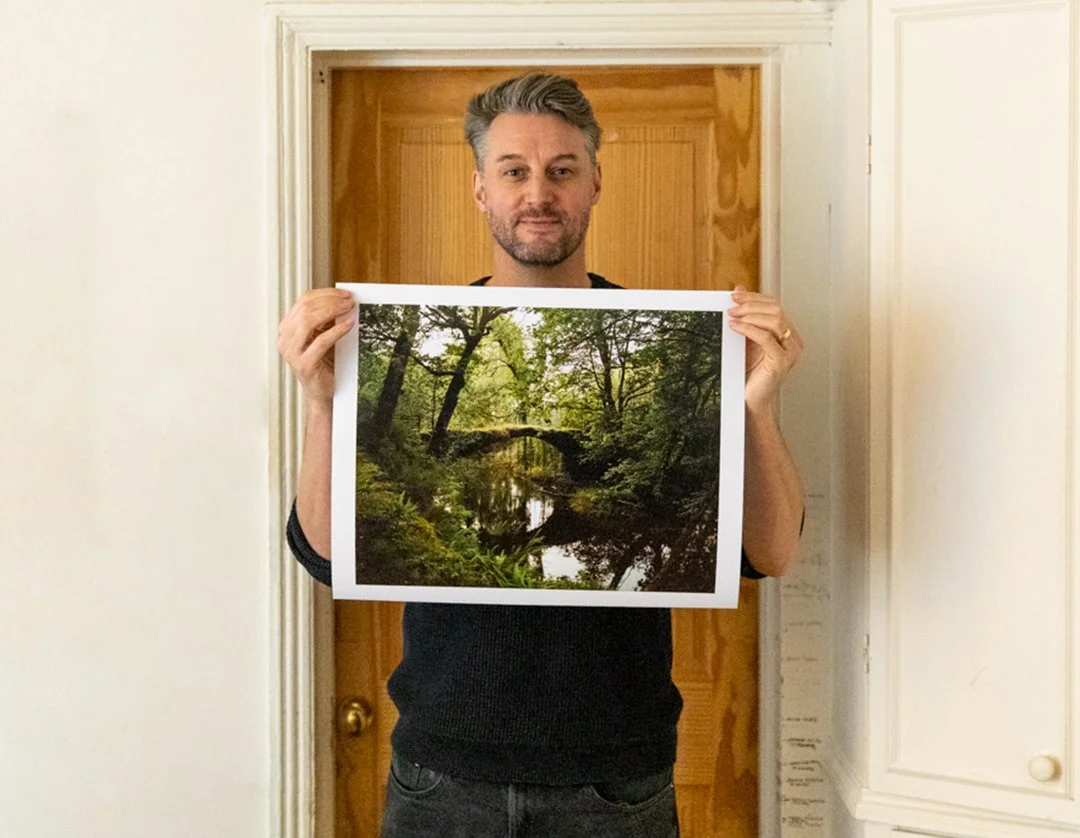 A man with gray hair and a beard holding a framed photograph of a lush, green forest with a small river or stream running through it, standing in front of a wooden door.