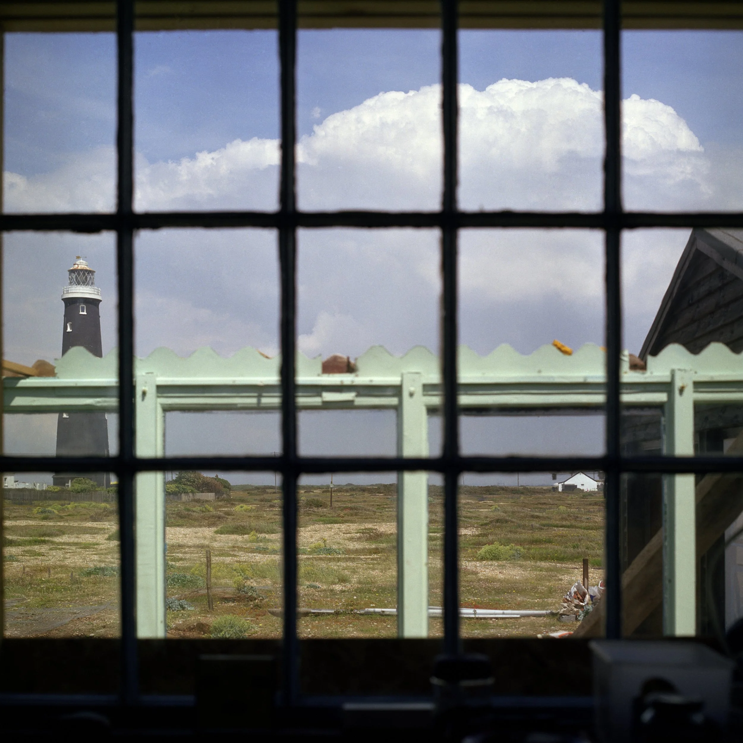 View through a window with black grid bars, showing a lighthouse on the coast and a cloudy sky in the background.