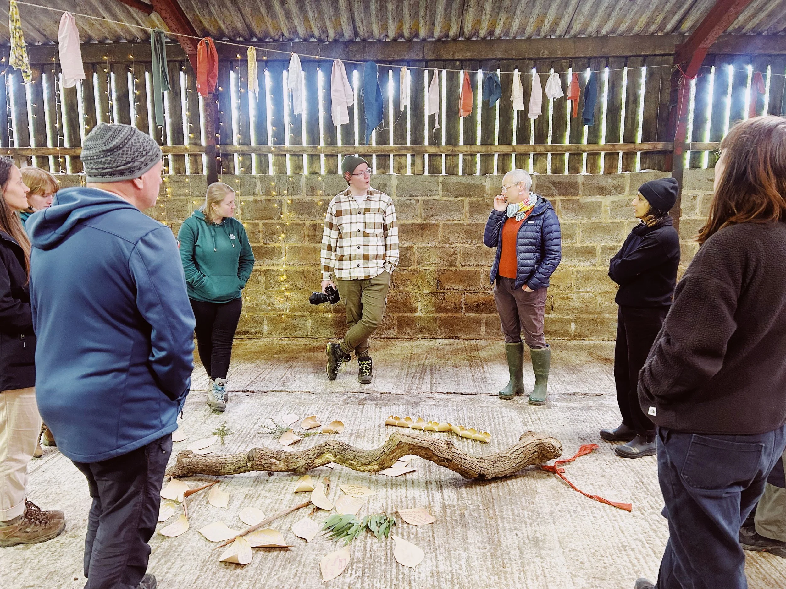 A group of people standing inside a rustic barn with wooden walls, string lights, and clothes hanging overhead. They are gathered around a display of natural items including leaves, a large branch, and animal bones on the ground.