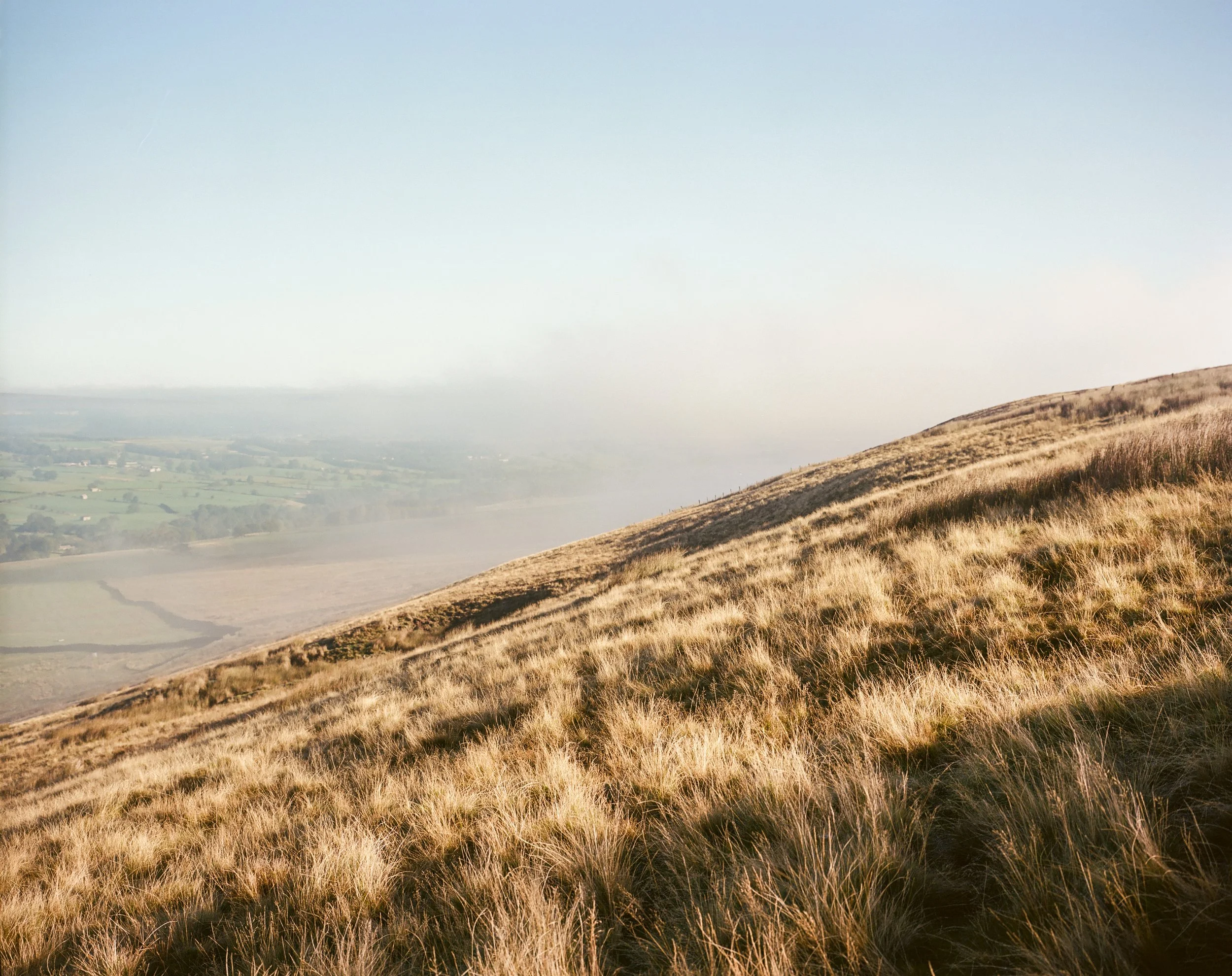 A hillside covered with dry yellowish grass during daytime, with a clear blue sky and a faint horizon line in the background.