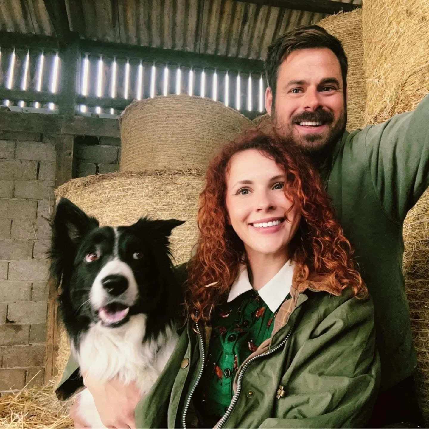 A smiling woman with curly red hair and a man with dark hair and beard taking a selfie in a barn with hay bales and a black and white dog.