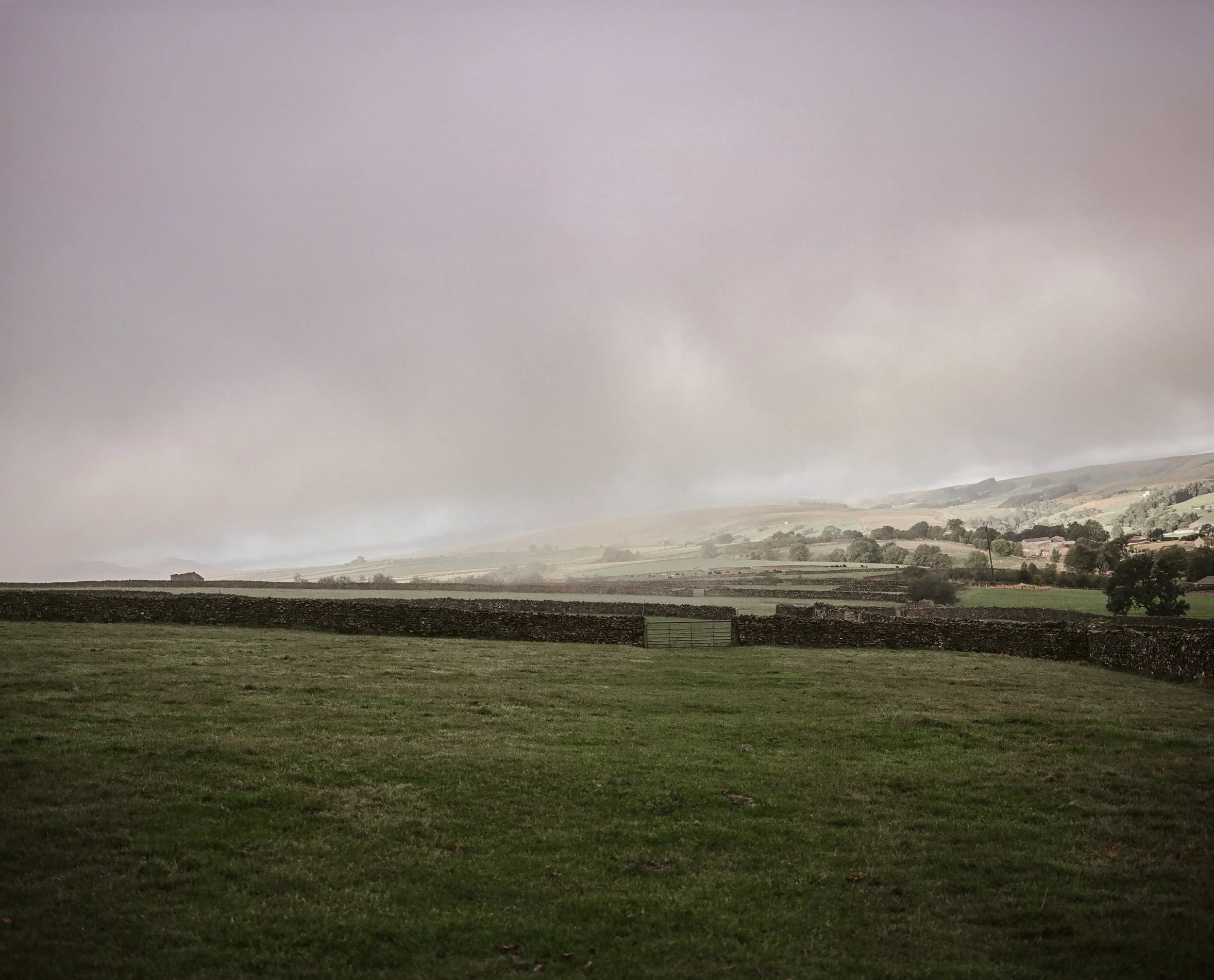 A landscape scene with a grassy field in the foreground, stone walls, rolling hills, and a cloudy sky.