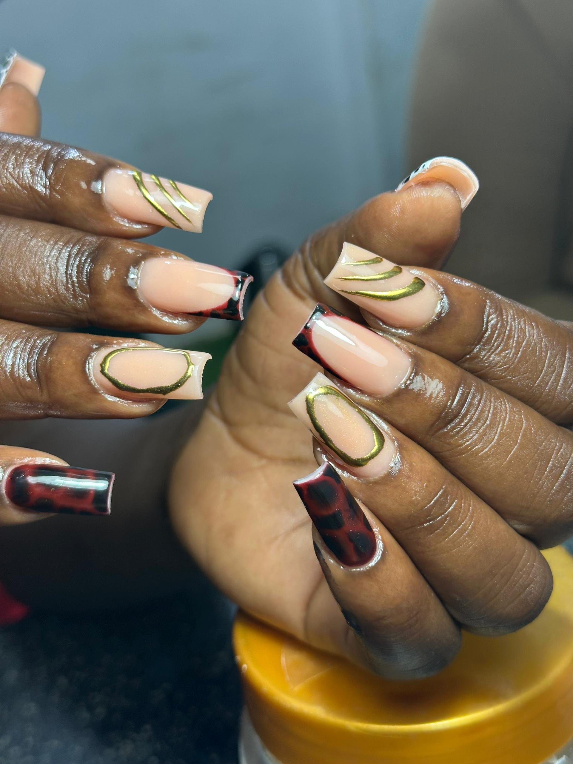 Close-up of a person's hands with intricately designed acrylic nails, featuring a mix of cream, red, black, green, and gold accents in various patterns.