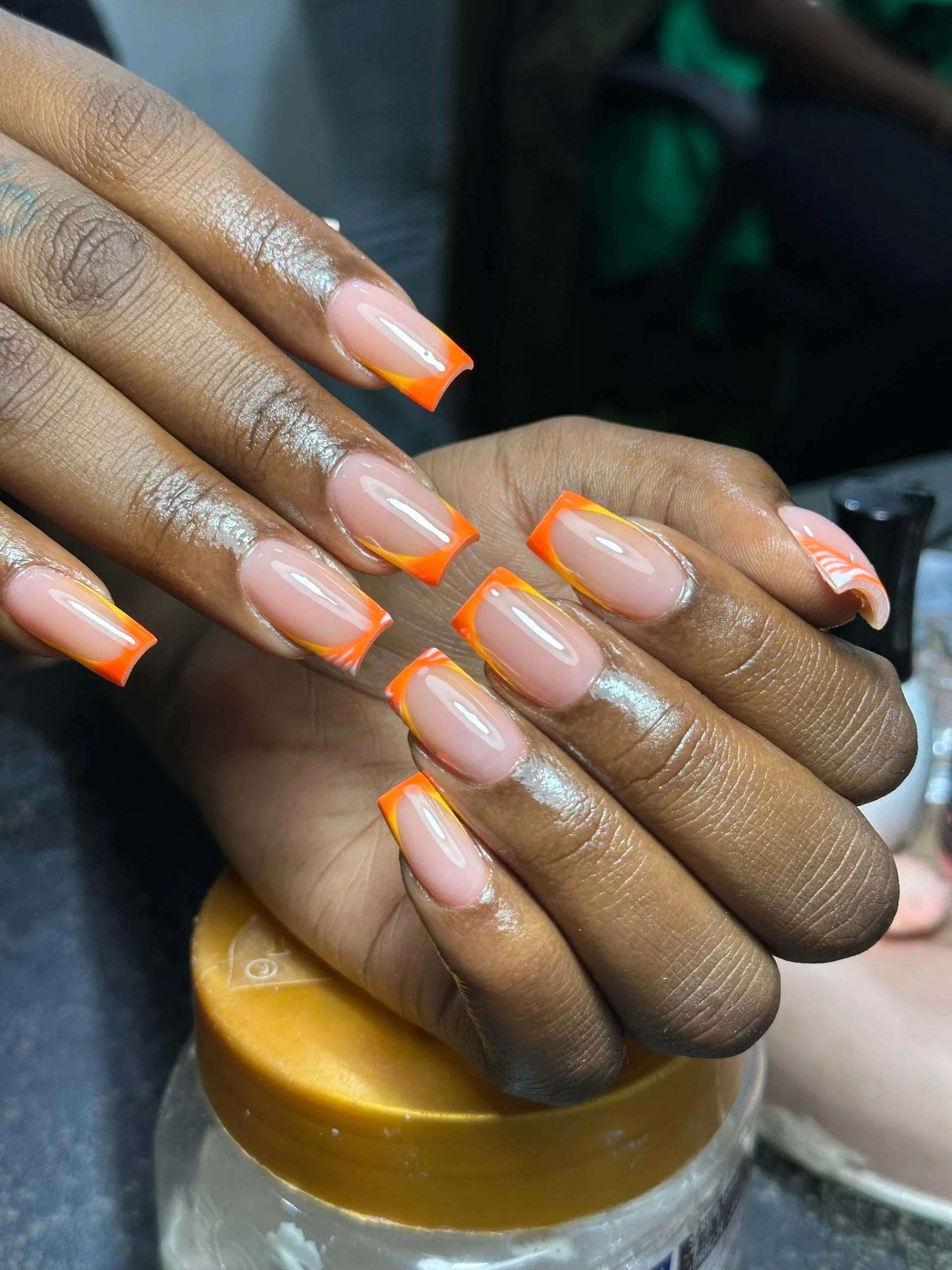 Close-up of a person's hands with freshly manicured nails featuring a clear base and orange tips with yellow accents, resting on a container with a golden lid.