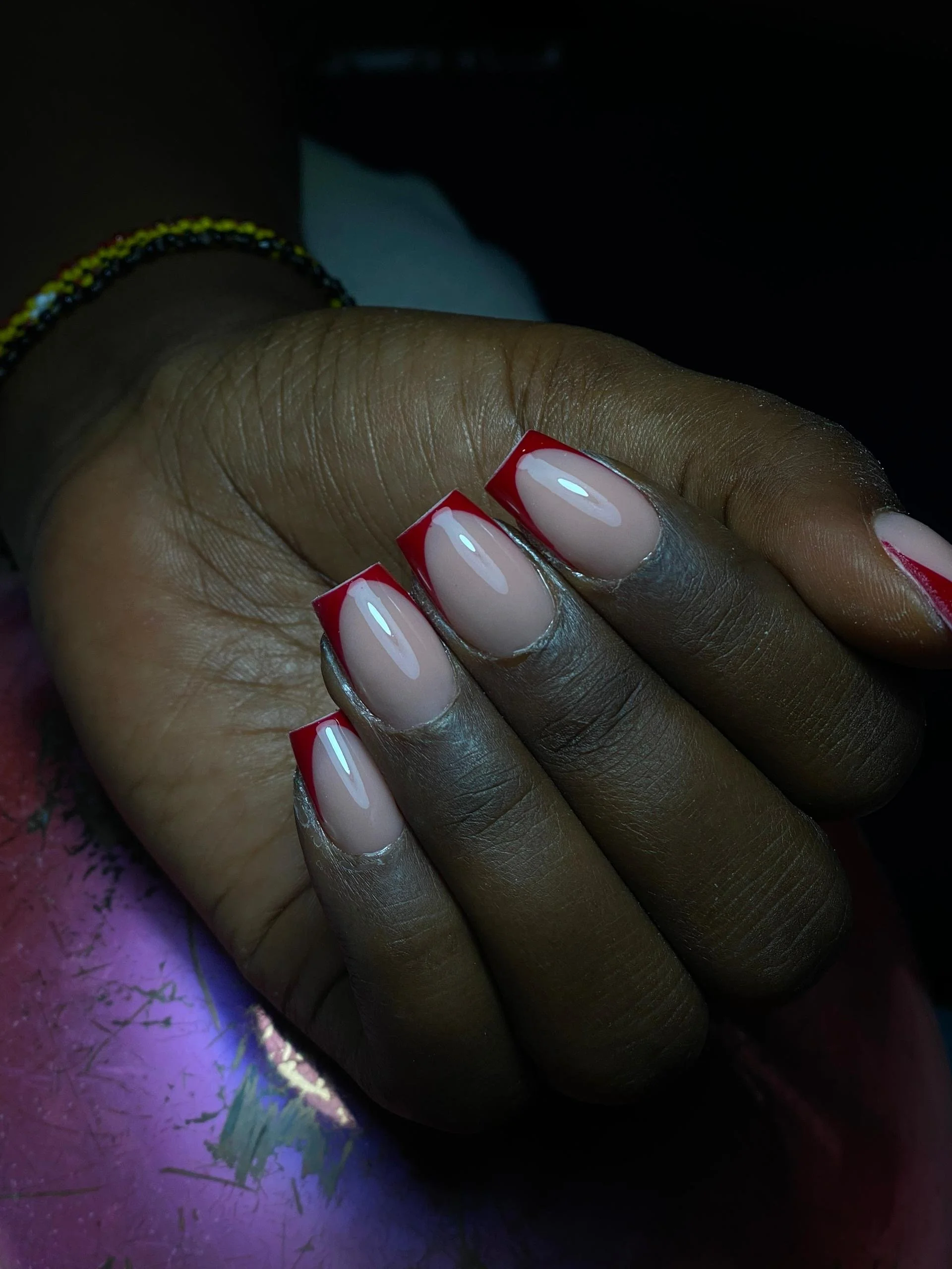 Close-up of a person's hand showcasing neatly manicured nails with red French tips and a natural base, resting on a colorful surface with dark background lighting.