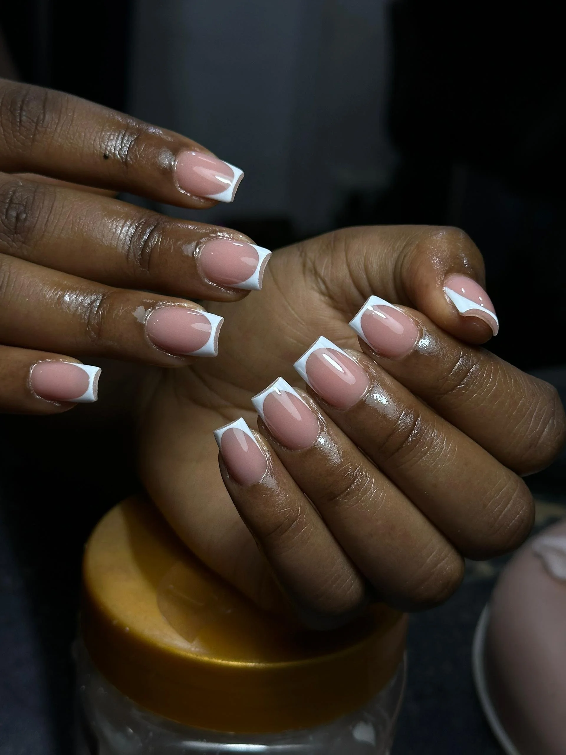 Close-up of a person's hands with manicured nails featuring a French tip design, positioned over a jar with a gold lid.