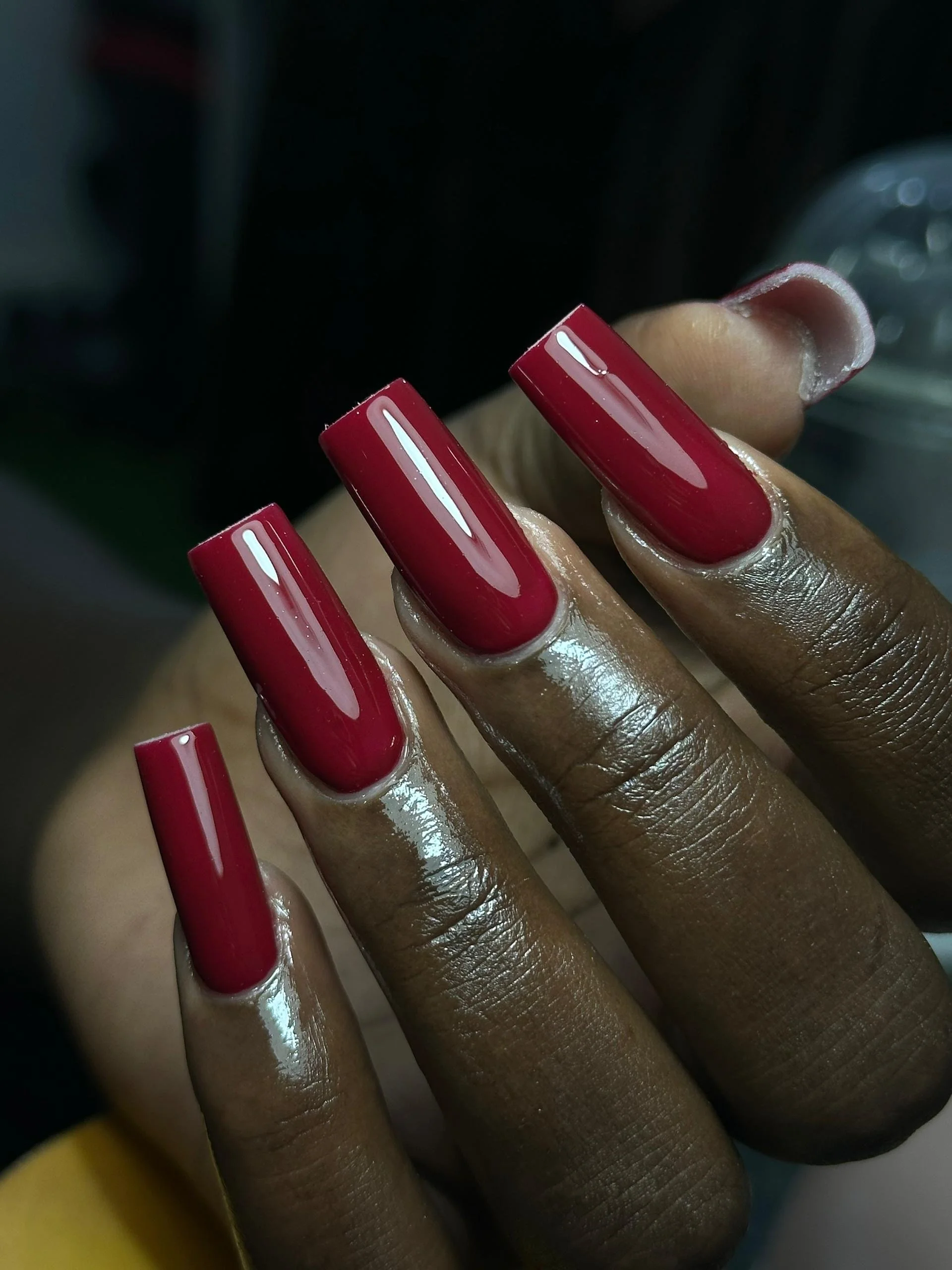Close-up of a hand with four fingers visible, showcasing long, glossy, deep red acrylic nails. The fingers are positioned to display the nails prominently, with a blurred background.