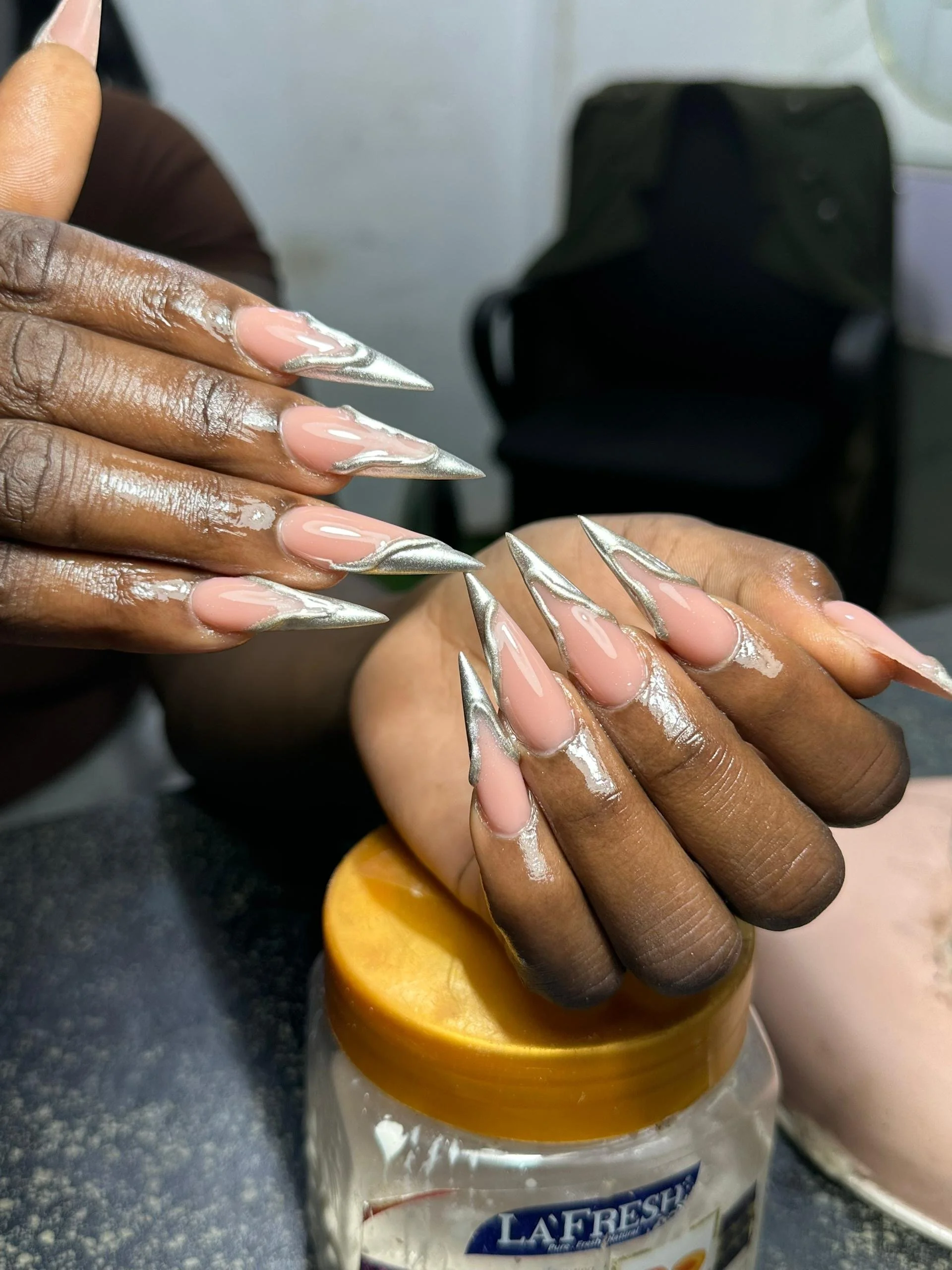 Close-up of a woman's hands with long, metallic silver stiletto nails, resting on a jar of La Fresh hand sanitizer or moisturizer in a salon or manicure setting.