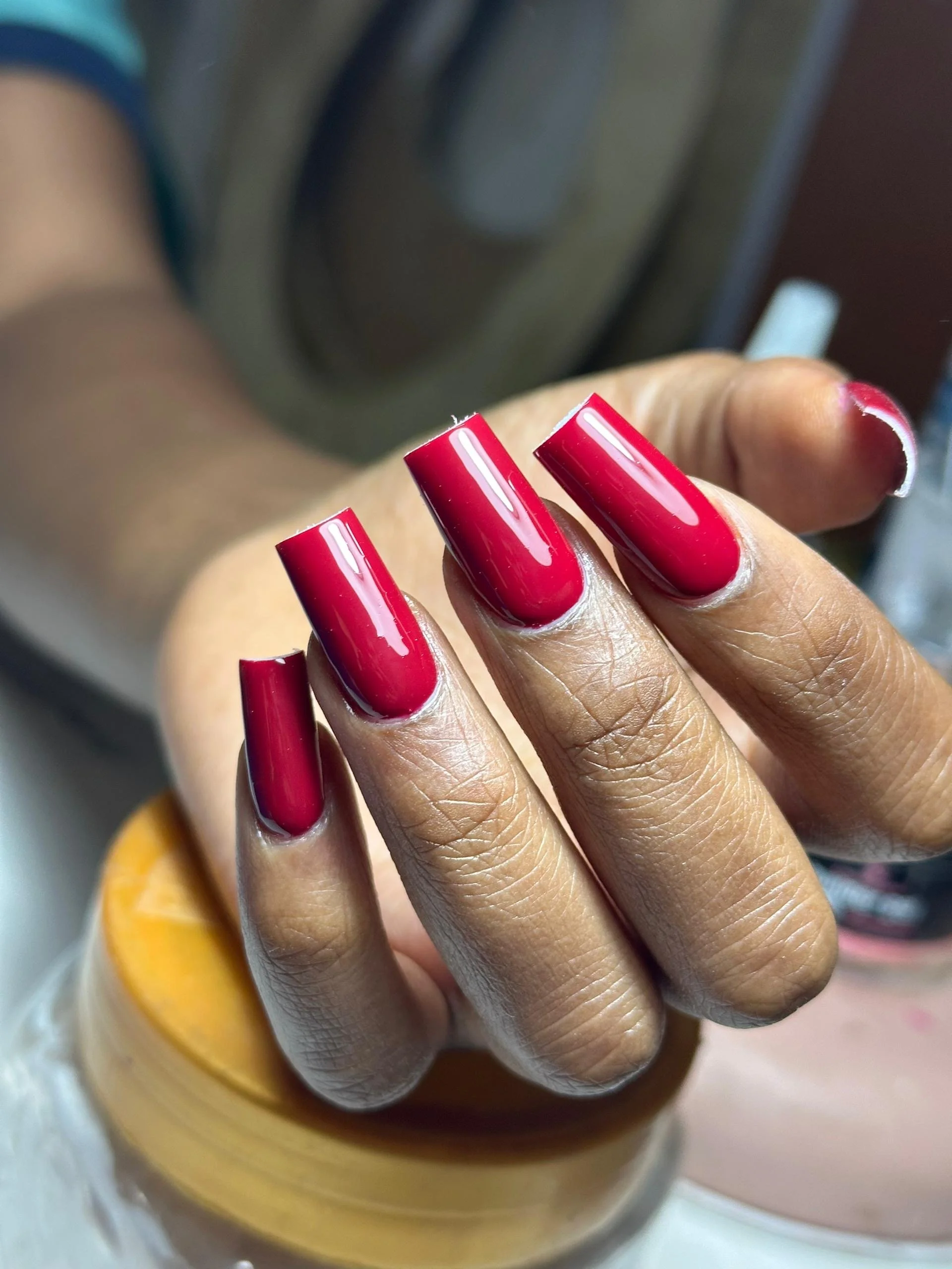 Close-up of a hand with freshly painted bright red, glossy, square-shaped nails. The background includes a blurred laundry machine.