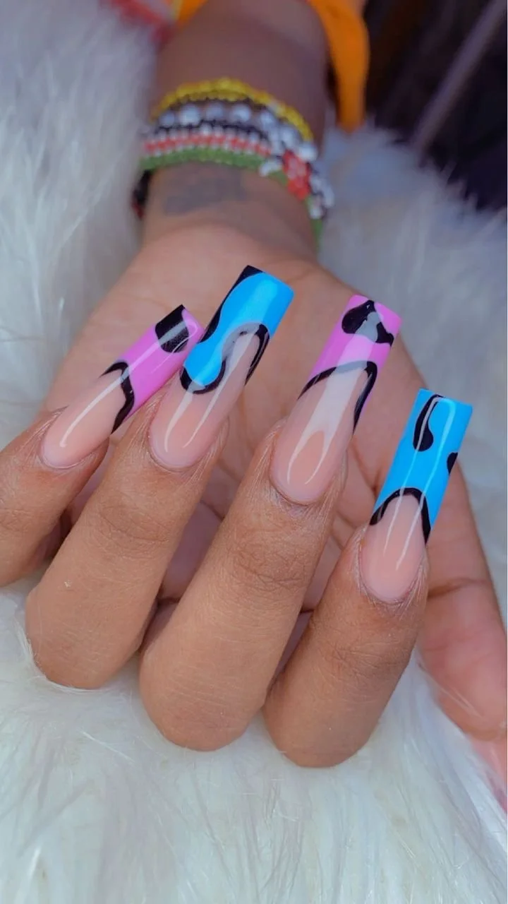 Close-up of a hand with long, colorful, marbled acrylic nails in pink, blue, black, and beige, resting on a soft white fur surface. In the background, a person's wrist is adorned with multicolored, beaded bracelets.