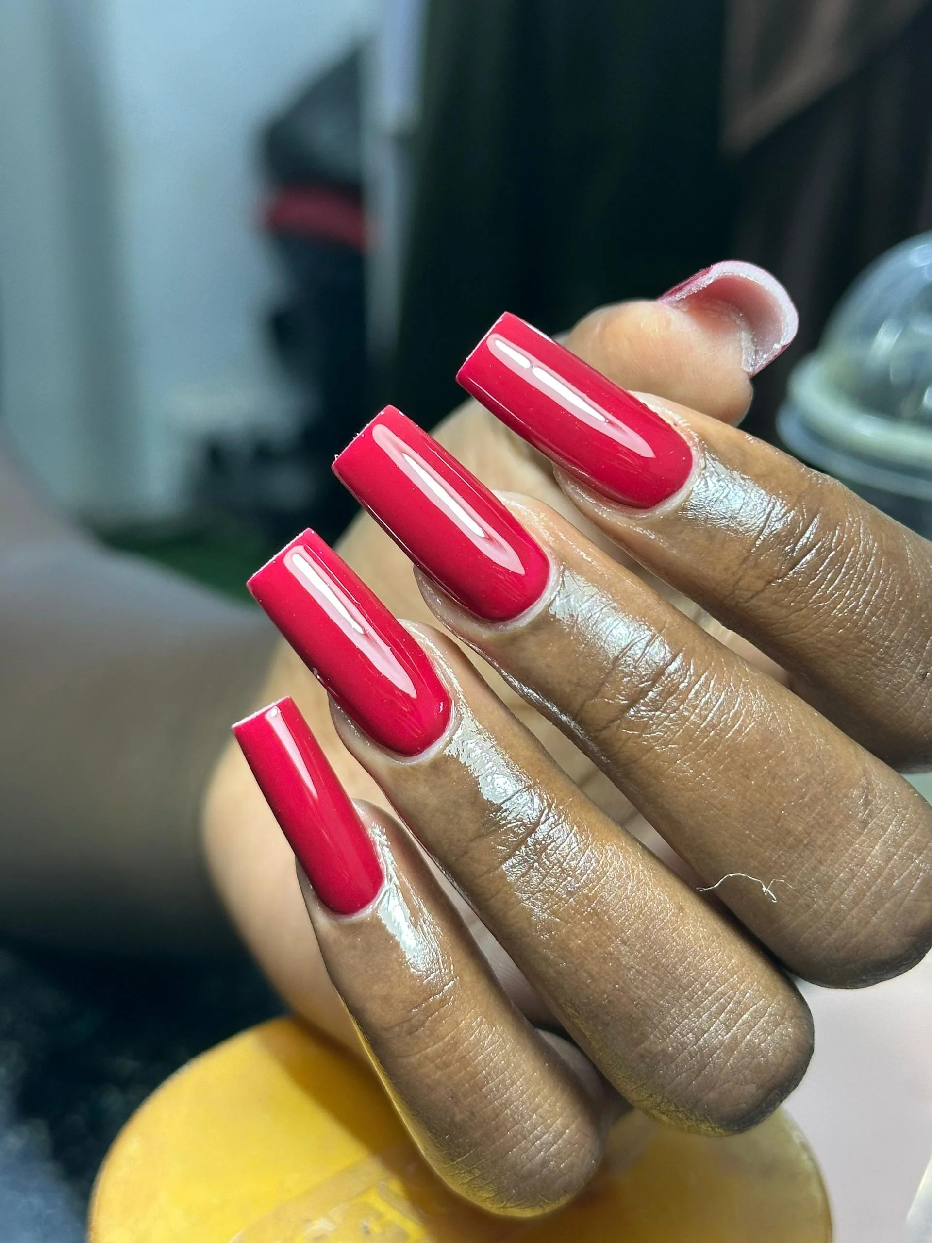 Close-up of a person's hand with long, shiny, bright red acrylic nails, with the fingers positioned across a yellow surface.
