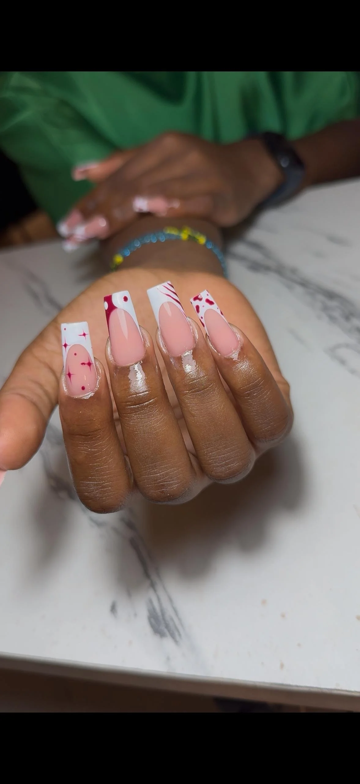 Close-up of a hand with decorated fingernails featuring red and white holiday-themed designs, with a person in a green shirt in the background resting their hands on a marble surface.