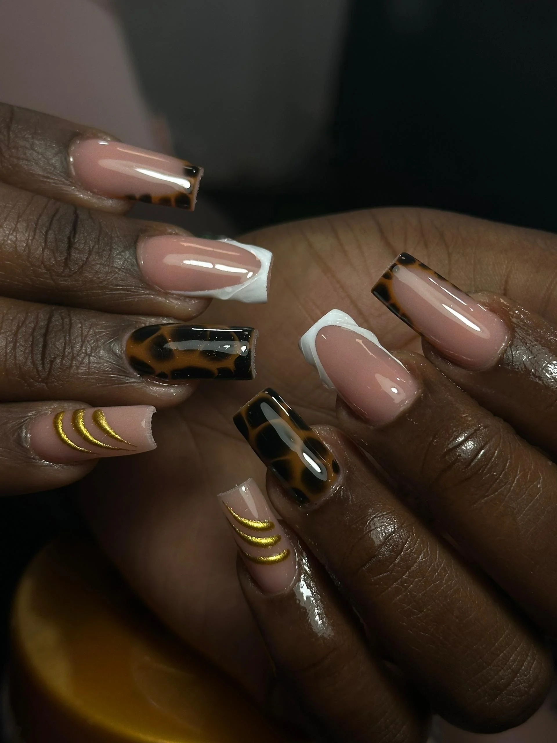 Close-up of a person’s hands with manicured nails featuring beige, white, and tortoiseshell designs, with some nails decorated with gold crescent stripes.