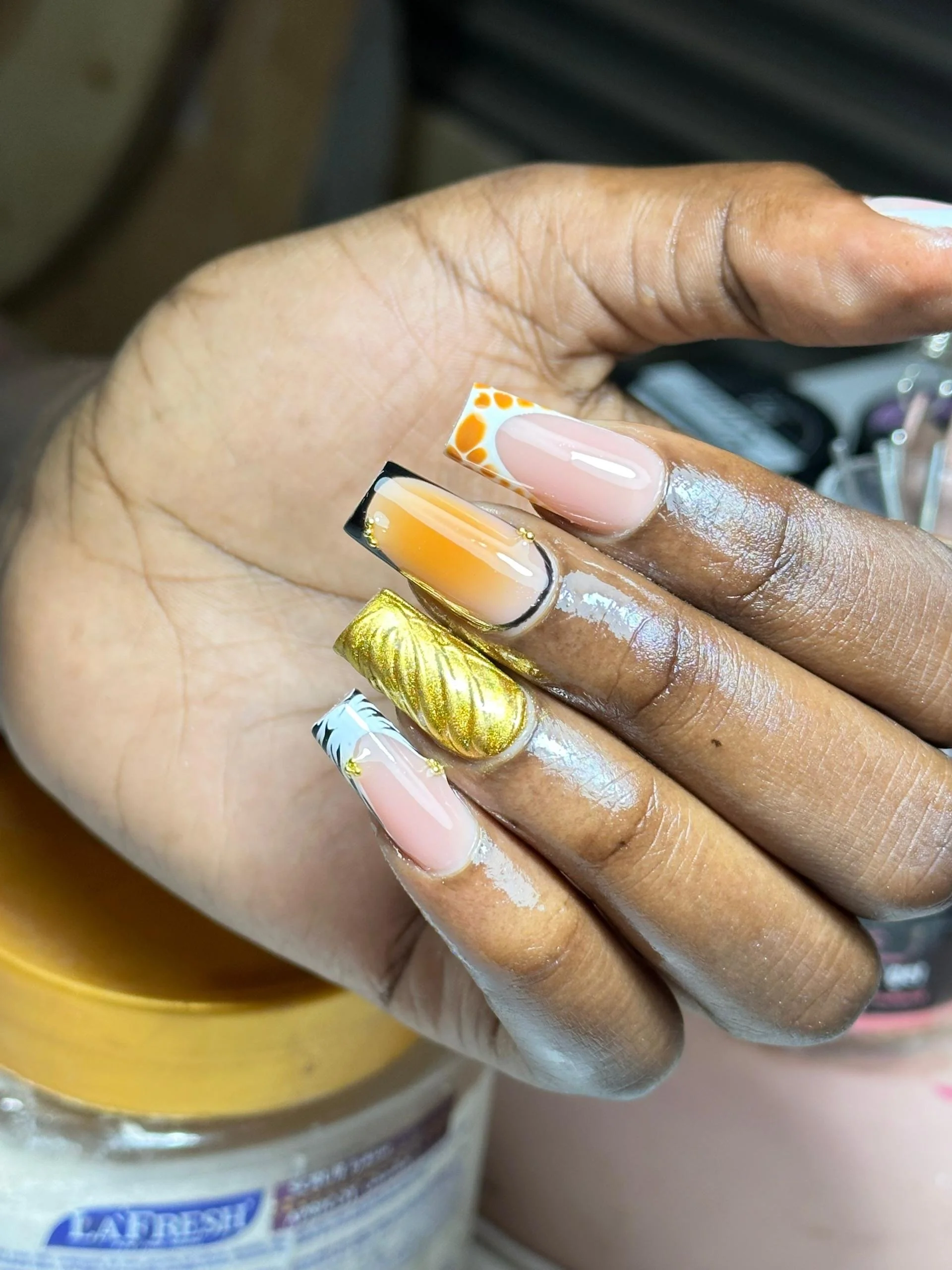 Close-up of a hand with decorated acrylic nails featuring various designs and colors, including leopard print, gradient, gold foil, and floral patterns.